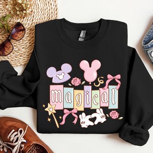May include: Black sweatshirt with the word "magical" in pastel colors, featuring Mickey Mouse ears, a pink balloon, roses, bows, a magic wand, and two white rabbits. The sweatshirt is displayed with sunglasses, shoes, and jeans.