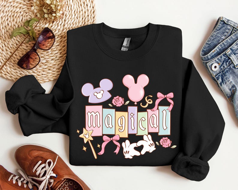 May include: Black sweatshirt with the word "magical" in pastel colors, featuring Mickey Mouse ears, a pink balloon, roses, bows, a magic wand, and two white rabbits. The sweatshirt is displayed with sunglasses, shoes, and jeans.