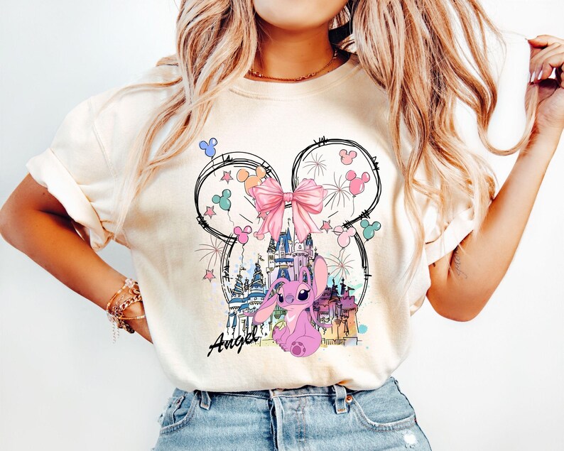 May include: Cream-colored t-shirt featuring a whimsical design. The graphic includes a pink bow, fireworks, and a castle within a Mickey Mouse ear outline. A pink cartoon character is seated below the castle, with the name "Angel" written in script.