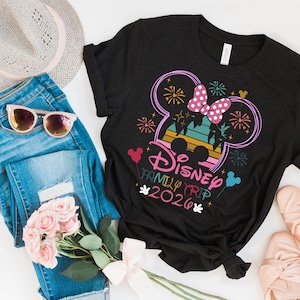 May include: Black t-shirt with a colorful Disney Family Trip 2020 graphic. The design includes a Mickey Mouse silhouette with a pink bow, fireworks, and the text "Disney Family Trip 2020". The shirt is paired with blue jeans, sunglasses, a hat, and pink shoes.