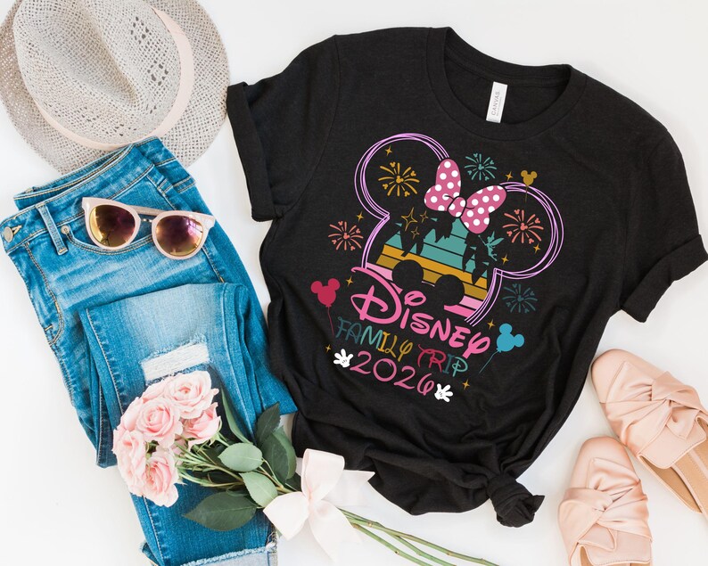 May include: Black t-shirt with a colorful Disney Family Trip 2020 graphic. The design includes a Mickey Mouse silhouette with a pink bow, fireworks, and the text "Disney Family Trip 2020". The shirt is paired with blue jeans, sunglasses, a hat, and pink shoes.
