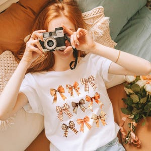 以下が含まれることがあります： 茶色とオレンジの格子縞のリボン模様の白いTシャツ。赤い髪の女性がヴィンテージカメラを持っている。