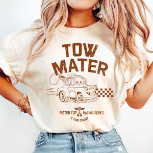 May include: Cream-colored t-shirt featuring a brown graphic of Tow Mater from the movie Cars. The text "TOW MATER" is above the cartoon image, with "Piston Cup Racing Series" and "5 Time Champ" below.