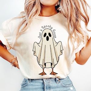 May include: Cream-colored t-shirt featuring a cartoon ghost with the text "Just a Spooky Goose". The ghost has black eyes and a beak, with orange feet. The shirt is a casual, everyday wear item.