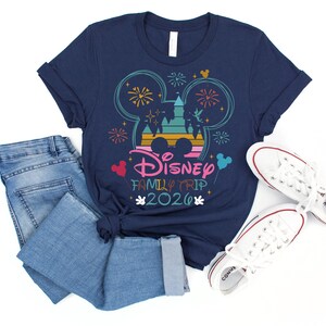 May include: Navy blue t-shirt with a colorful graphic featuring Mickey Mouse ears, fireworks, and a castle. The text "Disney Family Trip 2020" is displayed below the graphic. The shirt is paired with blue jeans and white sneakers.