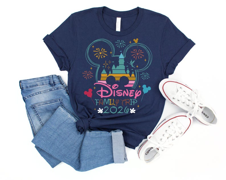 May include: Navy blue t-shirt with a colorful graphic featuring Mickey Mouse ears, fireworks, and a castle. The text "Disney Family Trip 2020" is displayed below the graphic. The shirt is paired with blue jeans and white sneakers.