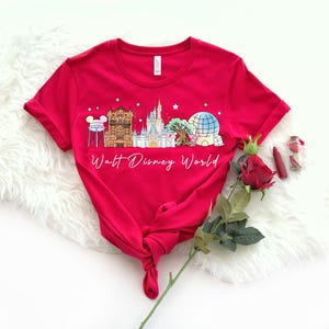 May include: A red t-shirt featuring a colorful graphic of Walt Disney World landmarks, including a castle, Spaceship Earth, and other attractions. The words "Walt Disney World" are written in a script font below the graphic.