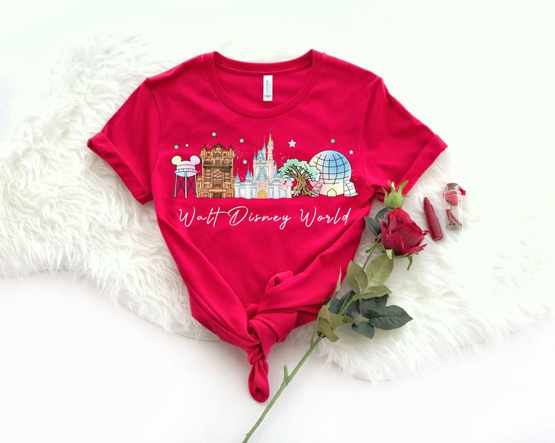 May include: A red t-shirt featuring a colorful graphic of Walt Disney World landmarks, including a castle, Spaceship Earth, and other attractions. The words "Walt Disney World" are written in a script font below the graphic.