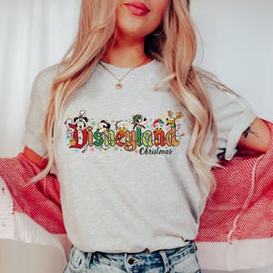 May include: A light gray t-shirt featuring the Disneyland Christmas logo with Mickey Mouse, Donald Duck, Goofy, and other characters. The design includes colorful text and festive holiday lights, perfect for a Disney fan.