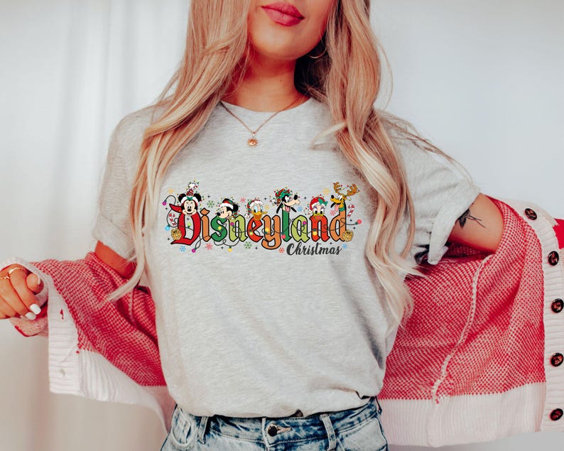 May include: A light gray t-shirt featuring the Disneyland Christmas logo with Mickey Mouse, Donald Duck, Goofy, and other characters. The design includes colorful text and festive holiday lights, perfect for a Disney fan.