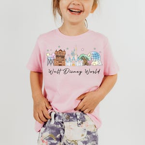May include: Pink t-shirt with a watercolor illustration of Walt Disney World landmarks. The shirt features the text "Walt Disney World" in a script font, perfect for a child.