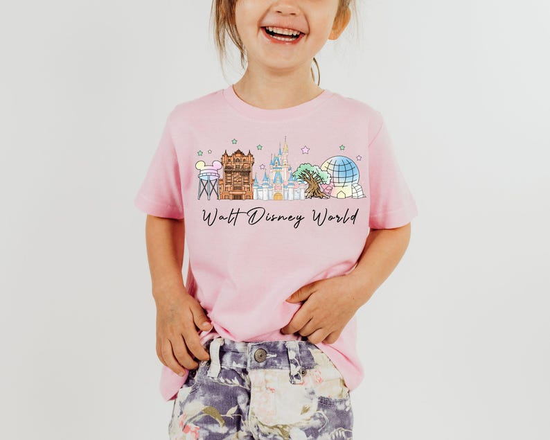 May include: Pink t-shirt with a watercolor illustration of Walt Disney World landmarks. The shirt features the text "Walt Disney World" in a script font, perfect for a child.