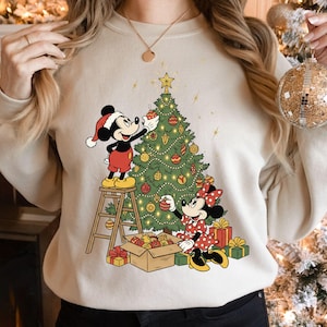 Puede incluir: Sudadera beige con una escena navideña. Mickey y Minnie Mouse decoran un árbol de Navidad. Mickey está en una escalera y Minnie en el suelo. El árbol está adornado con adornos y luces. Los regalos están al pie del árbol.