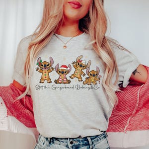 Puede incluir: Camiseta gris claro con un dise&ntilde;o de cuatro personajes Stitch de jengibre con decoraciones navide&ntilde;as. El texto "Stitch's Gingerbread Baking & Co" est&aacute; impreso debajo. La persona lleva una rebeca roja y blanca.
