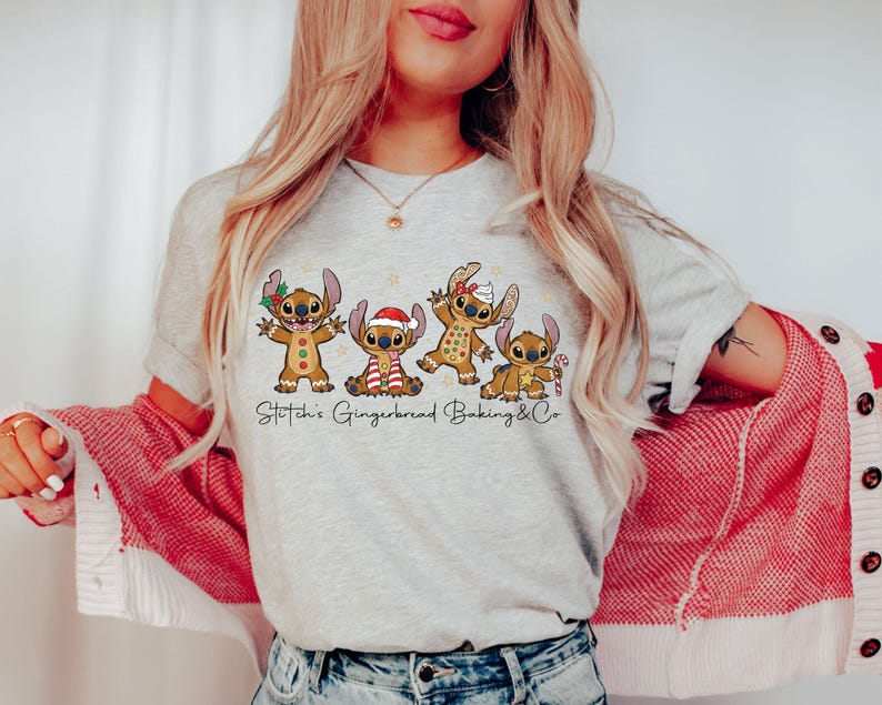 Puede incluir: Camiseta gris claro con un dise&ntilde;o de cuatro personajes Stitch de jengibre con decoraciones navide&ntilde;as. El texto "Stitch's Gingerbread Baking & Co" est&aacute; impreso debajo. La persona lleva una rebeca roja y blanca.