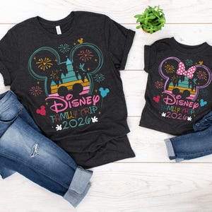 May include: Two dark gray t-shirts with a Disney Family Trip 2026 design, featuring Mickey Mouse ears, fireworks, and a castle silhouette. The shirts are paired with blue denim jeans. A small potted plant is also visible.