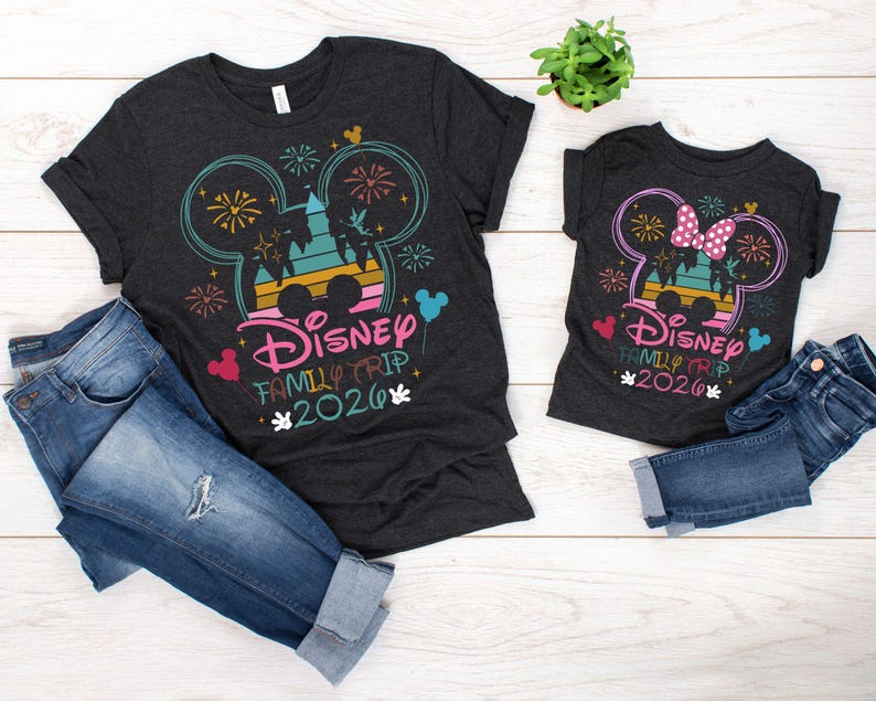 May include: Two dark gray t-shirts with a Disney Family Trip 2026 design, featuring Mickey Mouse ears, fireworks, and a castle silhouette. The shirts are paired with blue denim jeans. A small potted plant is also visible.