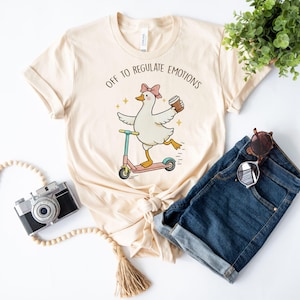 Puede incluir: Una camiseta de color crema con un gráfico de un ganso blanco con un lazo rosa, montado en un patinete rosa y sosteniendo una taza de café. El texto sobre el ganso dice OFF TO REGULATE EMOTIONS. El diseño incluye pequeñas estrellas amarillas.