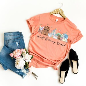 May include: Coral-colored t-shirt with a graphic of the Walt Disney World skyline, paired with blue jeans, black flats, and a bouquet of pink and white flowers. The shirt is hanging on a gold sequined hanger.