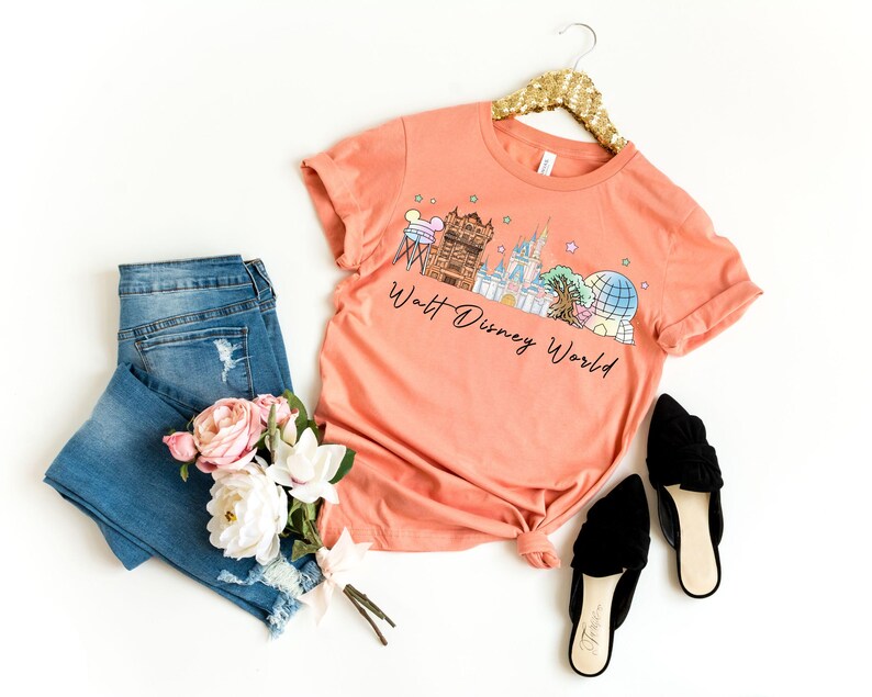 May include: Coral-colored t-shirt with a graphic of the Walt Disney World skyline, paired with blue jeans, black flats, and a bouquet of pink and white flowers. The shirt is hanging on a gold sequined hanger.