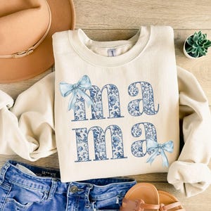 K&ouml;nnte beinhalten: Ein cremefarbener Pullover mit dem Wort "mama" in blauen und wei&szlig;en Blumenbuchstaben. Die Buchstaben sind schwarz umrandet und haben an jeder Seite eine blaue Schleife.