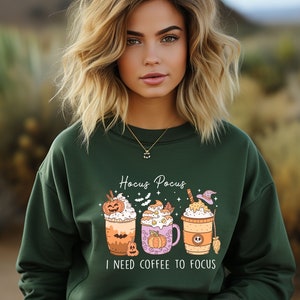 May include: A green sweatshirt with a Halloween-themed design featuring three coffee cups with pumpkin and witch hat decorations. The text "Hocus Pocus I need coffee to focus" is printed on the sweatshirt.