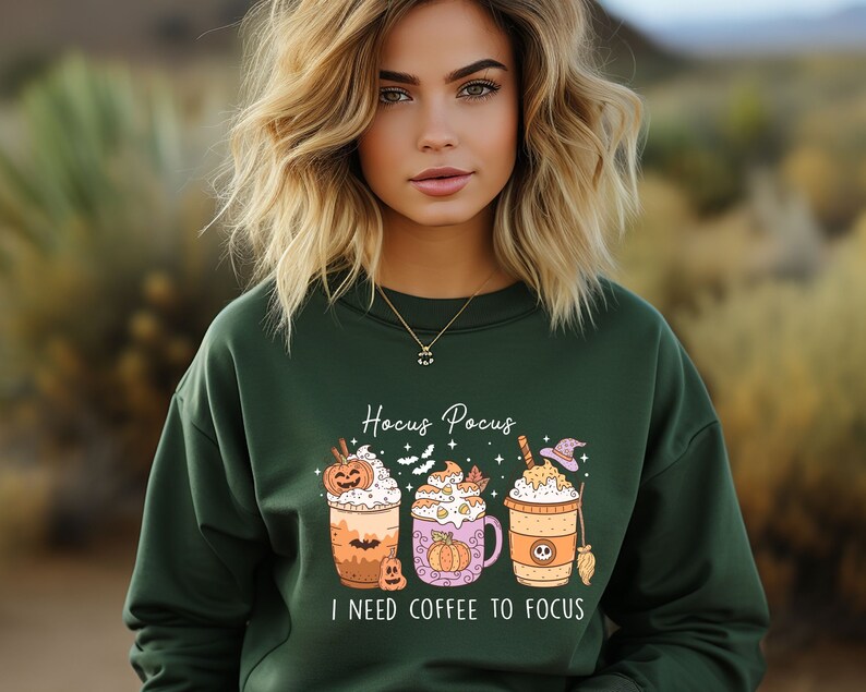 May include: A green sweatshirt with a Halloween-themed design featuring three coffee cups with pumpkin and witch hat decorations. The text "Hocus Pocus I need coffee to focus" is printed on the sweatshirt.
