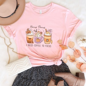 May include: Pink t-shirt with a graphic of three Halloween-themed coffee drinks and the text "Hocus Pocus I need coffee to focus".