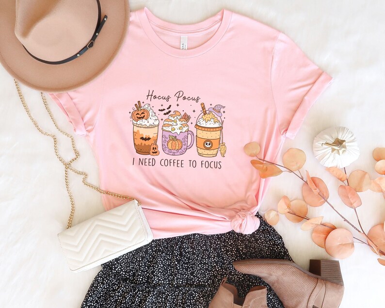 May include: Pink t-shirt with a graphic of three Halloween-themed coffee drinks and the text "Hocus Pocus I need coffee to focus".
