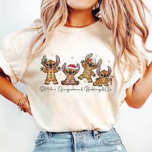 Puede incluir: Camiseta color crema con un gr&aacute;fico festivo de cuatro personajes Stitch vestidos de hombre de jengibre. El dise&ntilde;o incluye un gorro de Pap&aacute; Noel, un bast&oacute;n de caramelo y el texto "Stitch's Gingerbread Baking & Co". La camiseta es de material suave.
