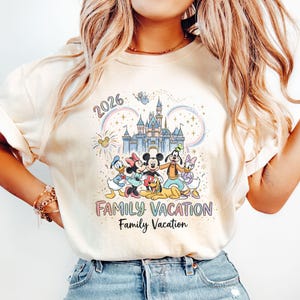 Może przedstawiać: Kremowa koszulka z kolorową grafiką postaci Disneya przed zamkiem. Projekt zawiera tekst "2026" i "Family Vacation". Koszulka ma krótkie rękawy i luźny krój.