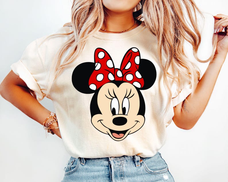 May include: A white t-shirt with a cartoon image of Minnie Mouse. Minnie Mouse is wearing a red bow with white polka dots and has a big smile.