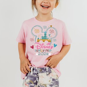 May include: A pink t-shirt featuring a colorful graphic of the Disney castle, fireworks, and Mickey Mouse ears. The text "Disney Family Trip 2020" is printed below the graphic. The shirt is worn by a young child.