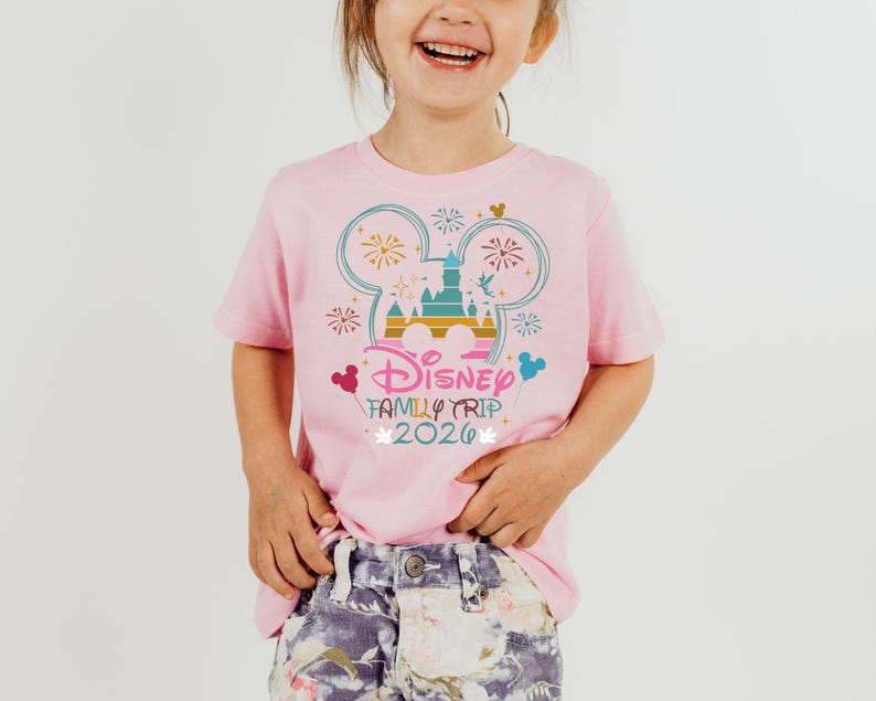May include: A pink t-shirt featuring a colorful graphic of the Disney castle, fireworks, and Mickey Mouse ears. The text "Disney Family Trip 2020" is printed below the graphic. The shirt is worn by a young child.