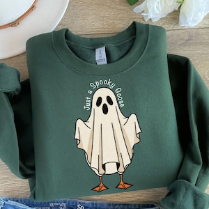 May include: A dark green sweatshirt with a ghost-goose graphic. The ghost is white with black eyes and a beak, and the text "Just a Spooky Goose" is printed above the ghost. The sweatshirt has a crew neck and long sleeves.