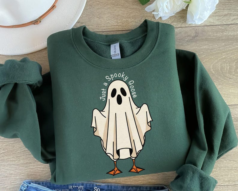 May include: A dark green sweatshirt with a ghost-goose graphic. The ghost is white with black eyes and a beak, and the text "Just a Spooky Goose" is printed above the ghost. The sweatshirt has a crew neck and long sleeves.