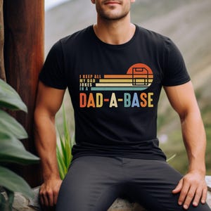 Könnte beinhalten: Schwarzes T-Shirt mit dem Text "I KEEP ALL MY DAD JOKES IN A DAD-A-BASE" in einem Retro-Design. Die Grafik zeigt einen Baseball-Backstop und ein Farbschema im Sonnenuntergangsstil in Orange, Gelb, Grün und Blau.