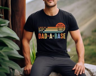 Op de afbeelding: Zwart t-shirt met de tekst "I KEEP ALL MY DAD JOKES IN A DAD-A-BASE" in retro-stijl. De afbeelding bevat een honkbal backstop en een kleurenschema in zonsondergangstijl van oranje, geel, groen en blauw.