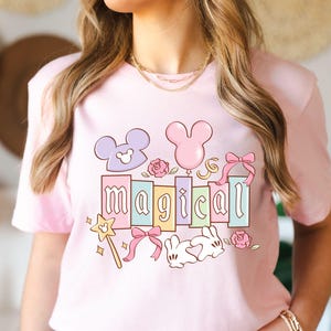 May include: A light pink t-shirt featuring a colorful graphic with the word "magical" in pastel colors. The design includes Mickey Mouse ears, a wand, bows, roses, and a bunny.