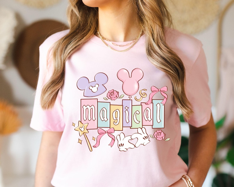 May include: A light pink t-shirt featuring a colorful graphic with the word "magical" in pastel colors. The design includes Mickey Mouse ears, a wand, bows, roses, and a bunny.