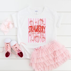 May include: White t-shirt with a red and pink strawberry graphic and the text "STRAWBERRY festival a berry good time." The image also includes a pink tutu skirt, pink trainers, and a pink bow.