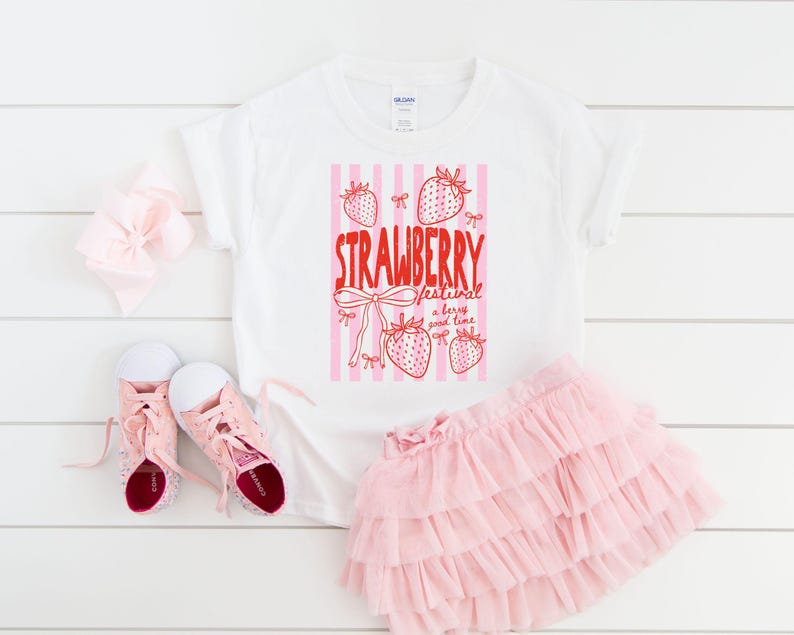 May include: White t-shirt with a red and pink strawberry graphic and the text "STRAWBERRY festival a berry good time." The image also includes a pink tutu skirt, pink trainers, and a pink bow.