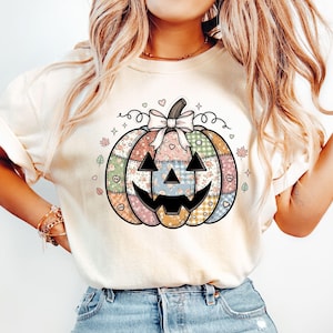 May include: Cream-colored t-shirt featuring a patchwork pumpkin design. The pumpkin has a smiling jack-o'-lantern face and is decorated with floral, plaid, and heart patterns. A bow and swirls adorn the top of the pumpkin.
