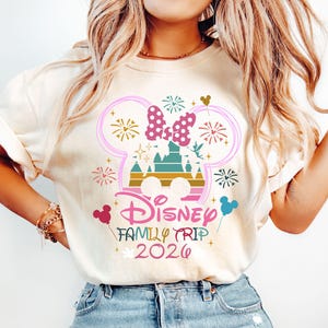 May include: Cream-colored t-shirt featuring a colorful graphic design. The design includes a castle, fireworks, and a bow, with the words "Disney Family Trip 2026". The design is in pink, teal, gold, and other colors.