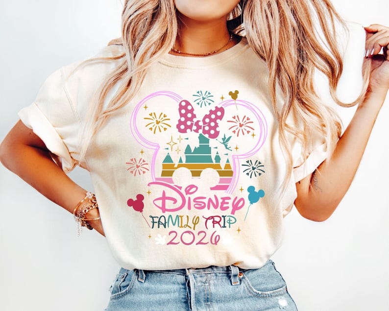 May include: Cream-colored t-shirt featuring a colorful graphic design. The design includes a castle, fireworks, and a bow, with the words "Disney Family Trip 2026". The design is in pink, teal, gold, and other colors.