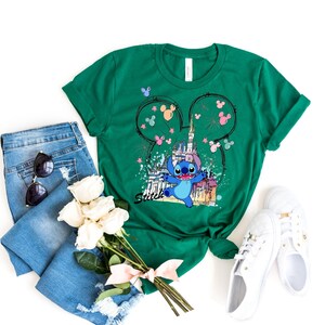 May include: Green t-shirt featuring a graphic of Stitch from Lilo & Stitch in front of a castle, with Mickey Mouse ears and the word "Smile". The shirt is styled with blue jeans, sunglasses, white sneakers, and a bouquet of white roses.