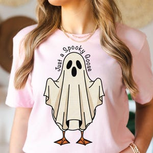 May include: A light pink t-shirt featuring a cartoon ghost with the text "Just a Spooky Goose". The ghost has black eyes and a beak, with orange feet. The design is centered on the shirt.
