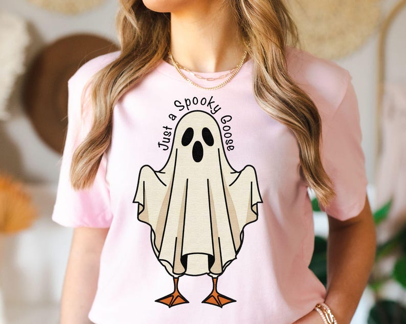 May include: A light pink t-shirt featuring a cartoon ghost with the text "Just a Spooky Goose". The ghost has black eyes and a beak, with orange feet. The design is centered on the shirt.