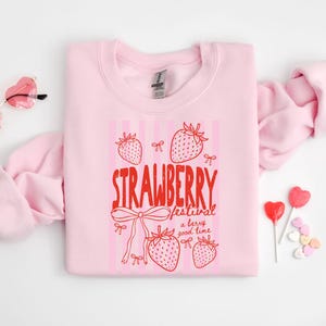 May include: A pale pink sweatshirt featuring the words "STRAWBERRY festival a berry good time" in red, with strawberry and bow illustrations. Heart-shaped sunglasses and sweets are also visible.
