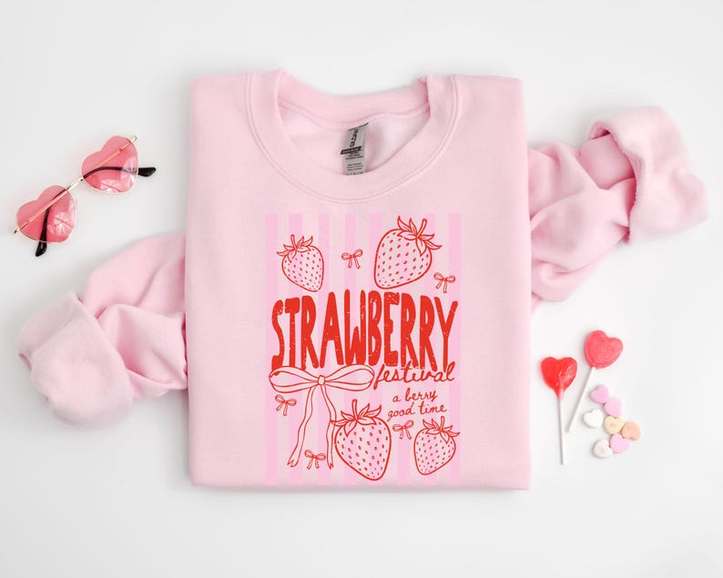 May include: A pale pink sweatshirt featuring the words "STRAWBERRY festival a berry good time" in red, with strawberry and bow illustrations. Heart-shaped sunglasses and sweets are also visible.
